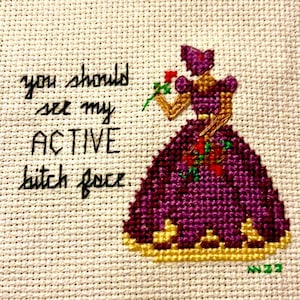 May include: Cross-stitch artwork on cream-coloured fabric. The text reads "you should see my ACTIVE bitch face." A purple dress with gold accents and a rose is depicted. The artwork is signed in the bottom right corner.