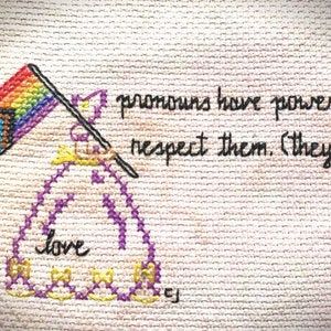 May include: A cross-stitch design with a rainbow flag and a purple heart with the word "love" inside. The text "pronouns have power. respect them. (they)" is stitched below the heart.