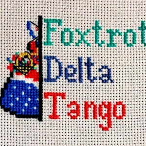 Peut inclure: Broderie au point de croix sur tissu blanc avec les mots "Foxtrot Delta Tango" en turquoise, bleu et rouge. Une image détaillée au point de croix d'une robe avec un panier de fleurs est sur le côté gauche.