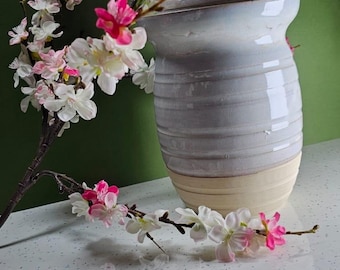 May include: A white ceramic jar with a lid and a light brown base. The jar is decorated with horizontal lines. The jar is sitting on a white countertop with a branch of white and pink flowers.