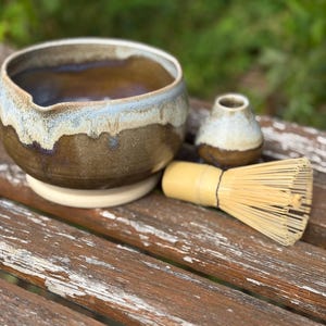 May include: A matcha tea set comprising a brown and cream ceramic bowl, a small ceramic tea cup, and a bamboo whisk. The bowl has a wide rim with a blue and cream glaze. The whisk is made of thin bamboo bristles.