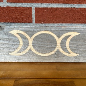 May include: A wooden sign with a triple moon symbol carved into it. The sign is painted gray with the symbol in a natural wood color.