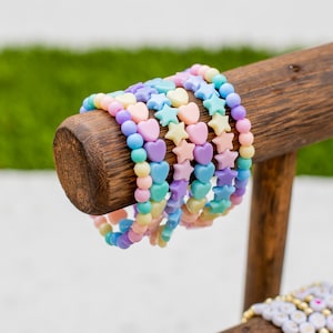 Könnte beinhalten: Pastellfarbene Armbänder mit Herz- und Sternanhängern werden auf einem Holzständer präsentiert. Die Armbänder weisen eine Vielzahl von sanften Farben auf, darunter Rosa, Gelb, Blau und Lila. Der Holzständer hat eine natürliche Oberfläche.