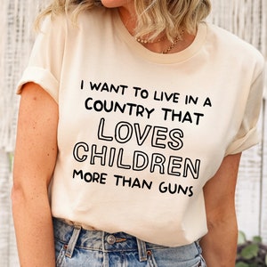 May include: A woman wearing a light beige t-shirt with the text "I want to live in a country that loves children more than guns" printed in black.