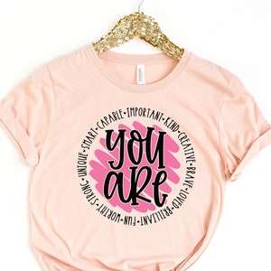 Puede incluir: Una camiseta rosa con un gráfico circular que dice "You are" en texto negro. El círculo está rodeado de una lista de palabras positivas en texto negro, incluyendo "smart", "capable", "important", "kind", "creative", "brave", "loved", "brilliant", "fun", "worthy", "strong", "unique".