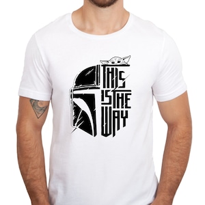 Puede incluir: Camiseta blanca con un gr&aacute;fico en blanco y negro de un casco mandaloriano y el texto "This is the way" con un peque&ntilde;o personaje asom&aacute;ndose por la parte superior del casco.