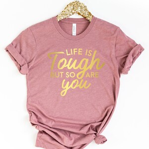 Puede incluir: Una camiseta color malva con el texto "Life is tough but so are you" en letras doradas.
