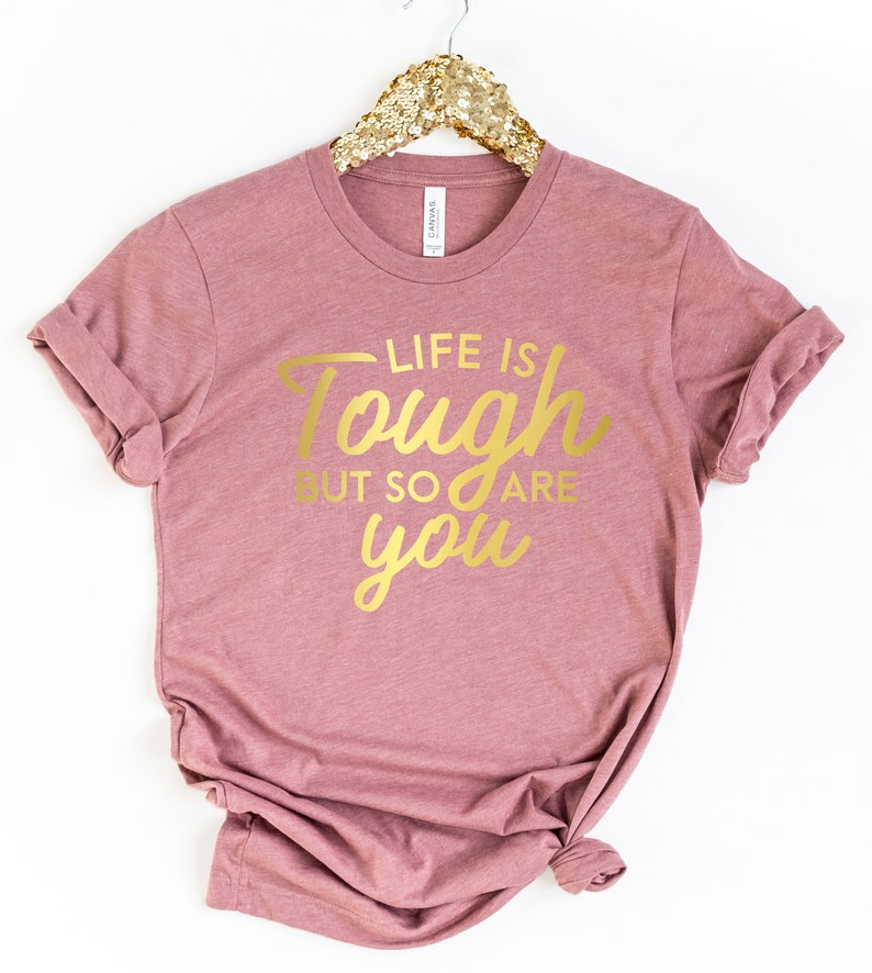 Puede incluir: Una camiseta color malva con el texto "Life is tough but so are you" en letras doradas.