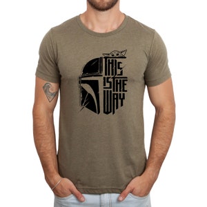 Puede incluir: Una camiseta verde con un gr&aacute;fico negro de un casco mandaloriano y el texto "This is the way" con un peque&ntilde;o personaje asom&aacute;ndose por la parte superior del casco.