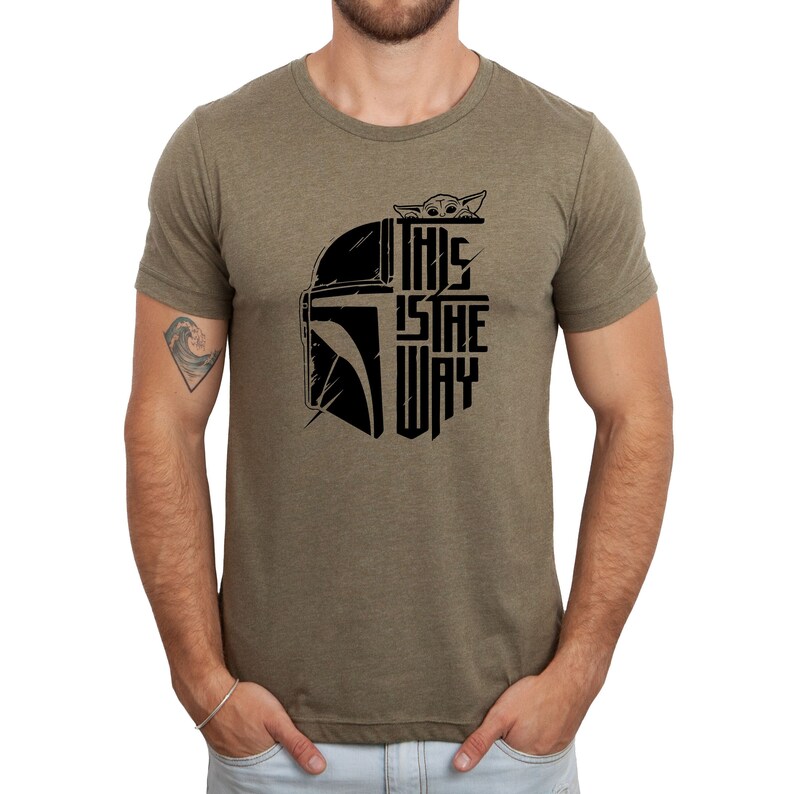 Puede incluir: Una camiseta verde con un gr&aacute;fico negro de un casco mandaloriano y el texto "This is the way" con un peque&ntilde;o personaje asom&aacute;ndose por la parte superior del casco.