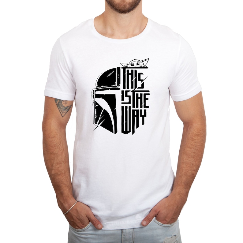 Puede incluir: Camiseta blanca con un gr&aacute;fico en blanco y negro de un casco mandaloriano y el texto "This is the way" con un peque&ntilde;o personaje asom&aacute;ndose por la parte superior del casco.