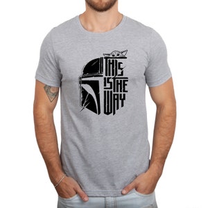 Puede incluir: Una camiseta gris con un gr&aacute;fico negro de un casco mandaloriano y el texto "This is the way" con un peque&ntilde;o personaje de Grogu asom&aacute;ndose por la parte superior del casco.