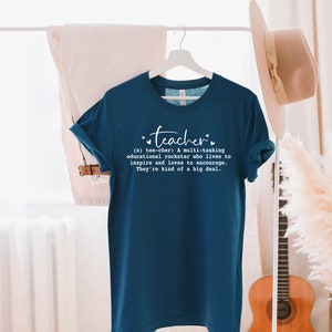 May include: A teal blue t-shirt with a white graphic that reads "teacher" and a definition of the word. The definition reads: "(n) tee-cher: A multi-tasking educational rockstar who lives to inspire and loves to encourage. They're kind of a big deal."