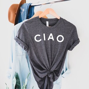 May include: Dark gray t-shirt with the word "CIAO" in white block letters. The shirt is knotted at the waist and hangs on a wooden hanger. Other clothing items and a hat are visible in the background.