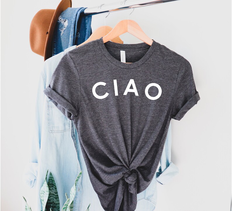 May include: Dark gray t-shirt with the word "CIAO" in white block letters. The shirt is knotted at the waist and hangs on a wooden hanger. Other clothing items and a hat are visible in the background.