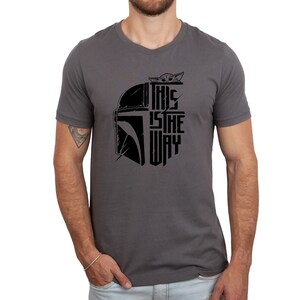 Puede incluir: Una camiseta gris oscuro con un gr&aacute;fico negro de un casco mandaloriano y el texto "This is the way" con una peque&ntilde;a imagen de Grogu asom&aacute;ndose por la parte superior del casco.