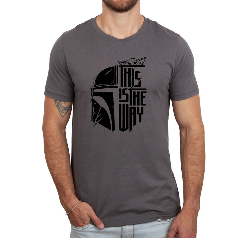 Puede incluir: Una camiseta gris oscuro con un gr&aacute;fico negro de un casco mandaloriano y el texto "This is the way" con una peque&ntilde;a imagen de Grogu asom&aacute;ndose por la parte superior del casco.
