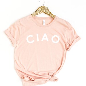 May include: A light pink t-shirt with the word "CIAO" printed in white. The shirt is hanging on a gold sequined hanger. The t-shirt is made of a soft material and has rolled-up sleeves.