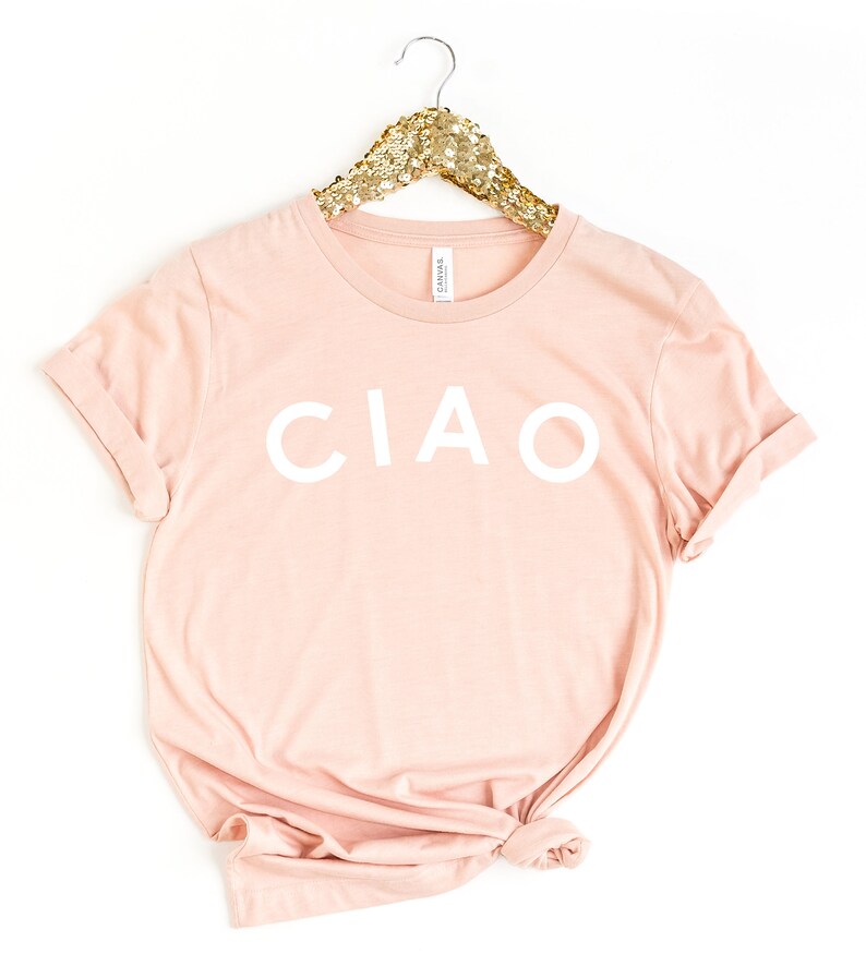 May include: A light pink t-shirt with the word "CIAO" printed in white. The shirt is hanging on a gold sequined hanger. The t-shirt is made of a soft material and has rolled-up sleeves.