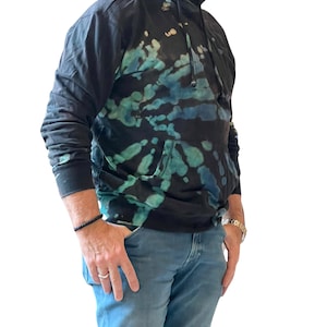 Może przedstawiać: Czarna bluza z kapturem z wzorem tie-dye w odcieniach turkusu i niebieskiego. Bluza ma kaptur ze sznurkiem, przednią kieszeń oraz prążkowane mankiety i dół. Osoba ma na sobie niebieskie dżinsy i bransoletkę.