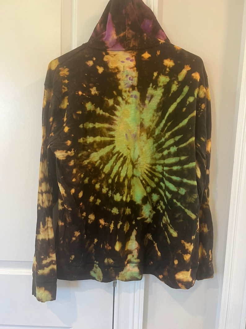 K&ouml;nnte beinhalten: Ein lang&auml;rmliger Kapuzenpullover mit einem Batik-Muster in den Farben Schwarz, Gr&uuml;n und Lila.