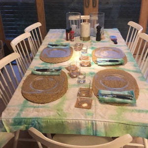Peut inclure: Une nappe blanche avec un motif tie-dye vert et bleu est dressée pour un dîner. La table est décorée de sets de table en osier, d'assiettes en verre transparent et d'argenterie. Il y a aussi des verres d'eau et des bougies sur la table.