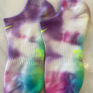 May include: A pair of white ankle socks with a tie-dye pattern in shades of purple, pink, blue, green, and yellow. The socks have a neon green Nike logo on the side.