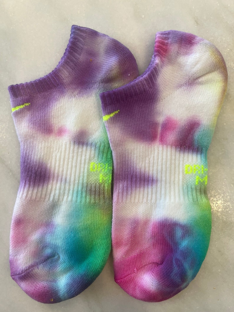 May include: A pair of white ankle socks with a tie-dye pattern in shades of purple, pink, blue, green, and yellow. The socks have a neon green Nike logo on the side.