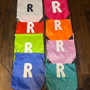 May include: Eight drawstring bags in different colours, each with a white letter "R" printed on the front. The bags are arranged in two rows of four, with the top row being white, pink, orange, and green, and the bottom row being red, light pink, blue, and teal.