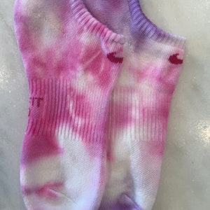 May include: A pair of white ankle socks with pink and purple tie-dye patterns. The socks have a small red logo on each sock.