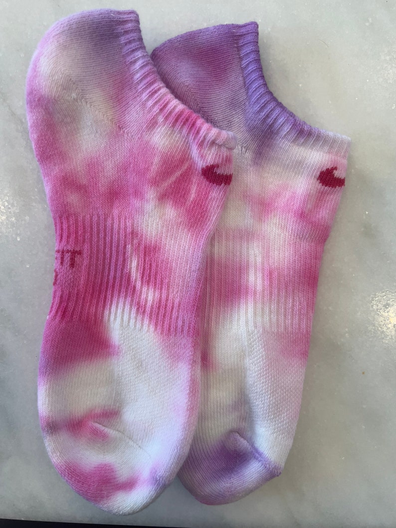 May include: A pair of white ankle socks with pink and purple tie-dye patterns. The socks have a small red logo on each sock.