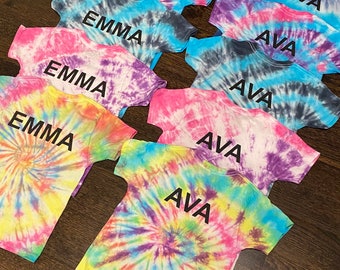 以下が含まれることがあります： 「Emma」と「Ava」の名前がプリントされたタイダイ染めのTシャツの積み重ね。Tシャツはピンク、ブルー、イエロー、パープルなど、さまざまな色があります。
