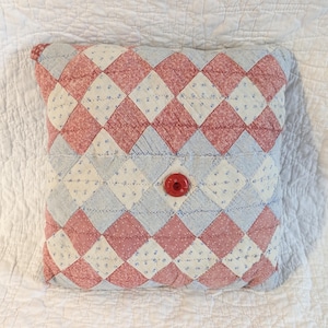 May include: Square quilted pillow with a patchwork design in red, white, and light blue diamond shapes. A large red button is centered on the pillow. The pillow is placed on a white quilted surface.
