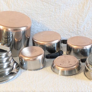 May include: A collection of stainless steel and copper-bottomed cookware, including pots, pans, and lids. The set features a variety of sizes, with black handles and a steamer insert. The copper bottoms provide a classic look for the kitchen.