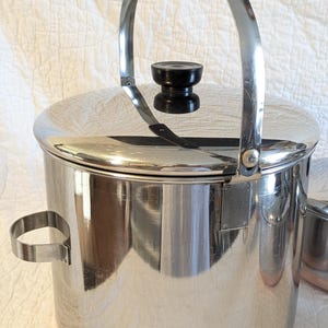May include: A stainless steel ice bucket with a black handle and lid knob. The bucket has a polished finish and a copper-coloured base. A small, matching cup is visible to the right.