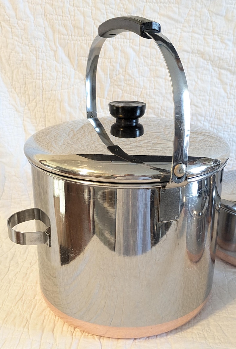 May include: A stainless steel ice bucket with a black handle and lid knob. The bucket has a polished finish and a copper-coloured base. A small, matching cup is visible to the right.