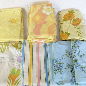 May include: A collection of folded vintage bed sheets with floral and striped patterns. The sheets feature various colors, including yellow, orange, pink, blue, and white. The floral designs include daisies and chrysanthemums.
