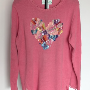 May include: A long-sleeved pink knit sweater featuring a heart-shaped design composed of multicolored sequined butterflies on the front. The neckline and hem have a scalloped edge, and a small sequined butterfly is located near the bottom left.