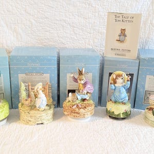May include: Five Beatrix Potter ceramic music boxes by Schmid, each featuring a different character scene on a circular base, accompanied by their original light blue boxes and a book titled The Tale of Tom Kitten by Beatrix Potter.