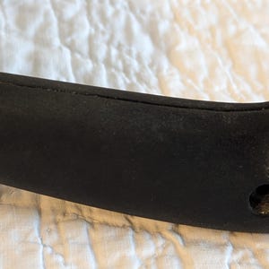 May include: Close-up of a black handle with a silver metal ring. The handle is made of a textured, dark material and is attached to a metal object. A screw is visible on the side of the handle.