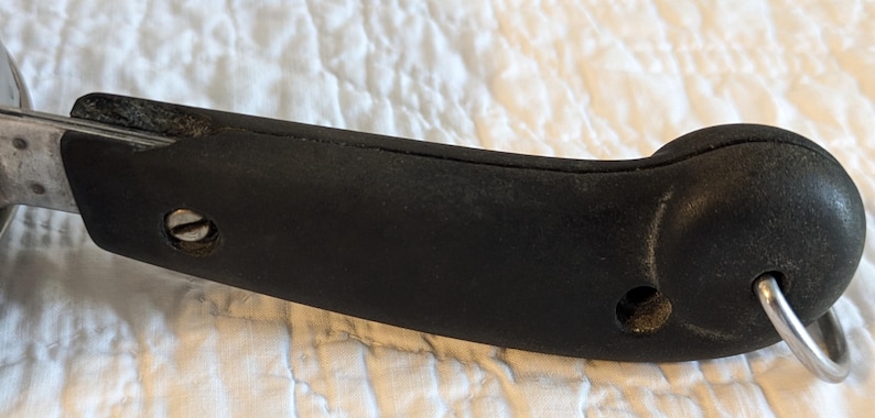 May include: Close-up of a black handle with a silver metal ring. The handle is made of a textured, dark material and is attached to a metal object. A screw is visible on the side of the handle.