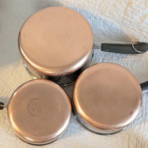 May include: Three copper-bottomed saucepans with black handles and silver wire stands. The copper bases have a shiny, reflective surface. The pans are arranged on a white, textured surface, showcasing their vintage design.