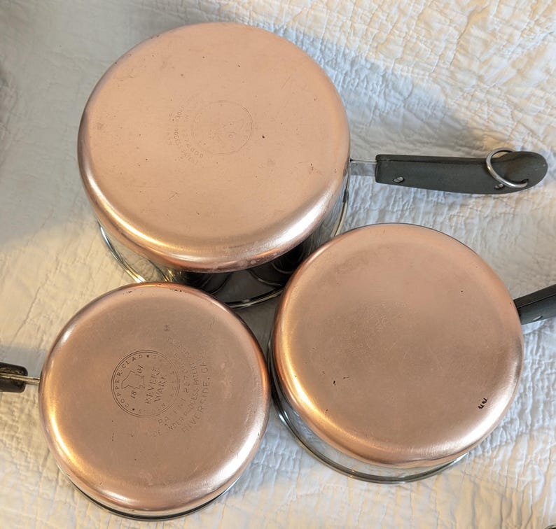 May include: Three copper-bottomed saucepans with black handles and silver wire stands. The copper bases have a shiny, reflective surface. The pans are arranged on a white, textured surface, showcasing their vintage design.