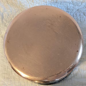 May include: A circular copper-coloured pot lid. The lid has a polished surface with some minor marks. The rim is a darker colour, possibly black. The overall appearance suggests a vintage or well-used kitchen item, suitable for cooking.
