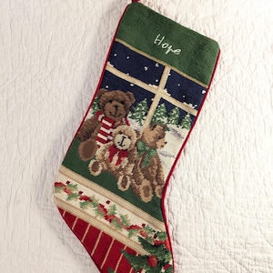 May include: A needlepoint Christmas stocking featuring three brown teddy bears sitting in front of a window with a snowy night scene. The stocking has a green cuff embroidered with the name Hope, a holly leaf border, and a red base with a tree.