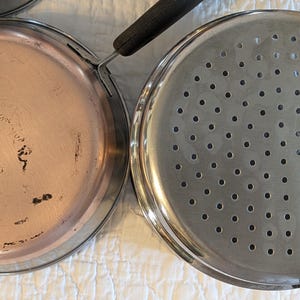 May include: Close-up of two stainless steel pans with handles. One pan has a copper-coloured bottom, and the other has many small holes. The pans are likely used for cooking and are placed on a white surface.