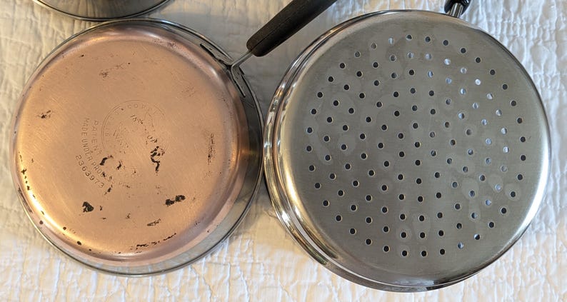 May include: Close-up of two stainless steel pans with handles. One pan has a copper-coloured bottom, and the other has many small holes. The pans are likely used for cooking and are placed on a white surface.