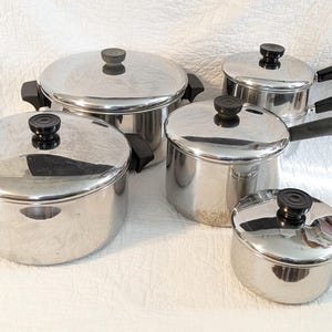 May include: A set of four stainless steel pots and pans with polished silver surfaces, black plastic handles, and matching lids. Each lid features a central black knob, and the cookware includes various sizes for cooking, all arranged together.