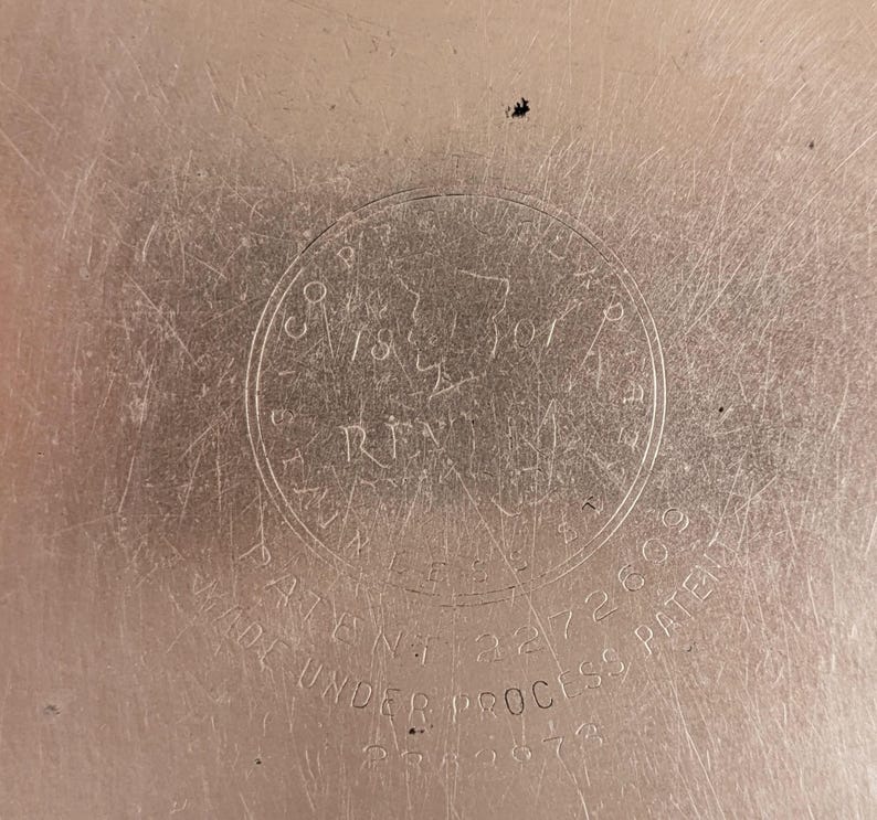 May include: Close-up of a metallic surface with a circular emblem. The emblem features text including "CO. 1870 RIVER" and "PATENT 2272609". The words "MADE UNDER PROCESS PATENT" are also visible. The surface has a brushed texture.