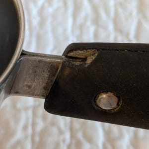 May include: Close-up of a metal and black handle attached to a stainless steel container. The handle is made of black material with a silver rivet. The metal shows signs of wear.